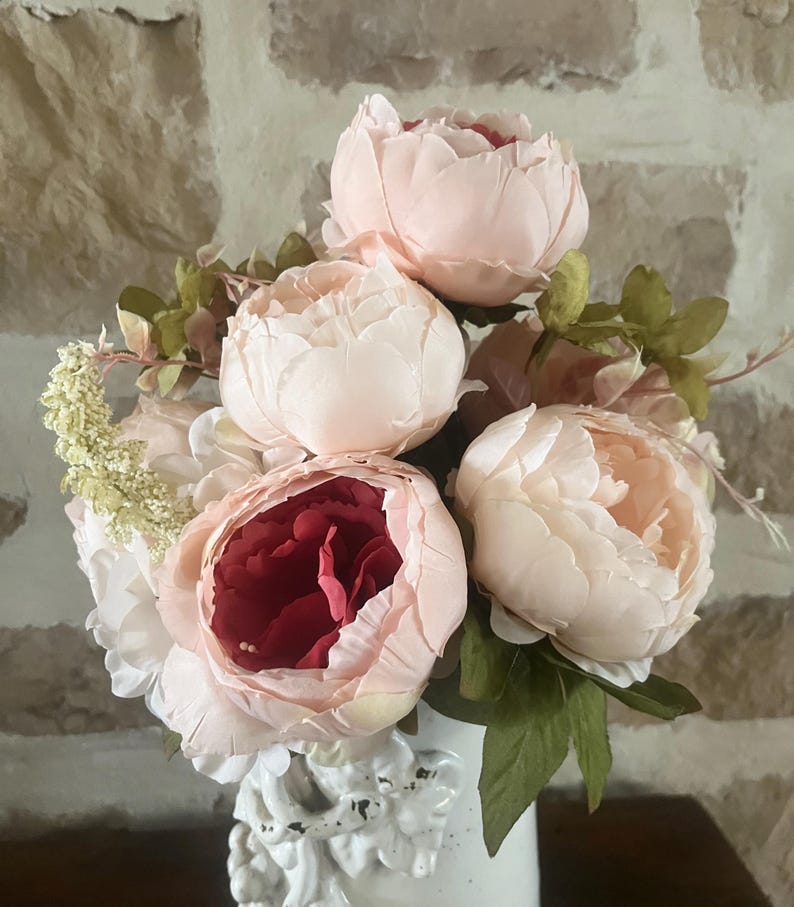 May include: A bouquet of pink and red peonies with green leaves in a white vase. The peonies are arranged in a cluster, with the pink peonies on top and the red peony in the center.