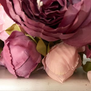 May include: Close-up of a bouquet of artificial flowers. The arrangement features large, fully bloomed peonies in shades of deep burgundy and soft pink, with smaller rose buds in varying pink hues. Green leaves are interspersed.