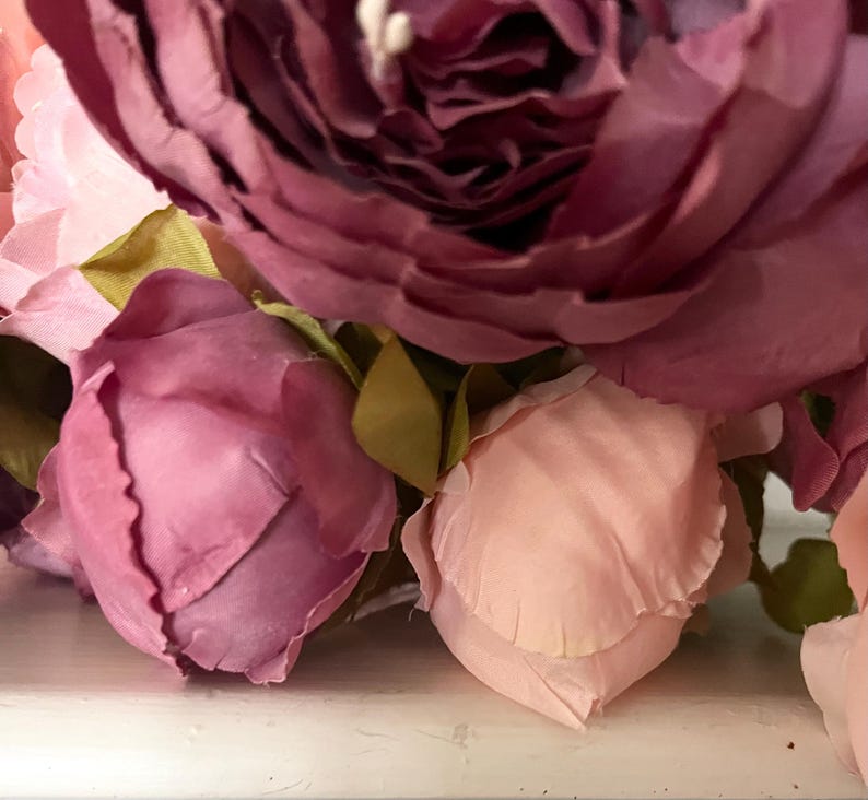 May include: Close-up of a bouquet of artificial flowers. The arrangement features large, fully bloomed peonies in shades of deep burgundy and soft pink, with smaller rose buds in varying pink hues. Green leaves are interspersed.