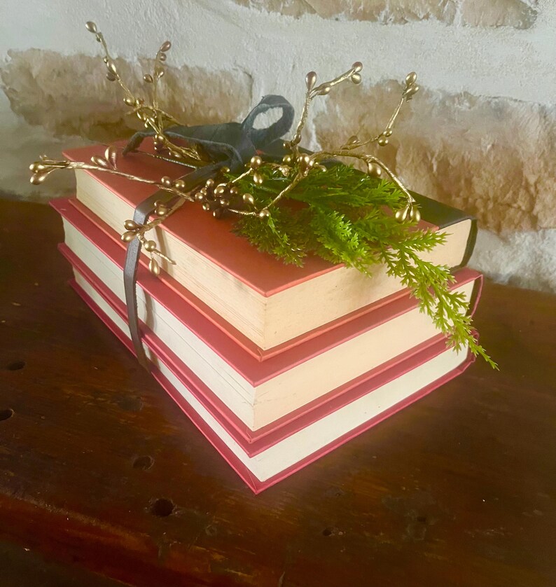 May include: A stack of three books with red covers and white pages, tied with a dark gray ribbon. The books are decorated with a sprig of green foliage and gold-colored decorative branches.