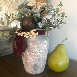 May include: A white ceramic vase with a raised design of Santa Claus holding a sack of gifts. The vase is decorated with a red velvet ribbon and a bouquet of faux greenery, silver ornaments, and pine cones.