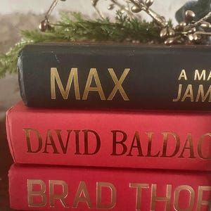 May include: A stack of three hardcover books. The top book is black with the gold-colored title "MAX". The middle and bottom books are red with gold lettering. Decorative greenery and gold-colored sprigs are in the background.