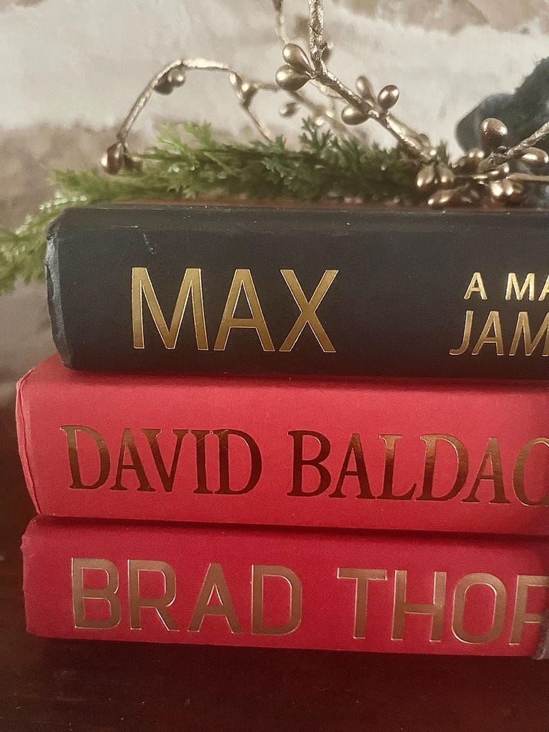 May include: A stack of three hardcover books. The top book is black with the gold-colored title "MAX". The middle and bottom books are red with gold lettering. Decorative greenery and gold-colored sprigs are in the background.