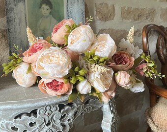May include: A floral arrangement featuring large, soft-colored peonies in shades of pink, peach, and white, accented with green foliage. The flowers are arranged on a decorative, ornate table. A framed portrait is in the background.