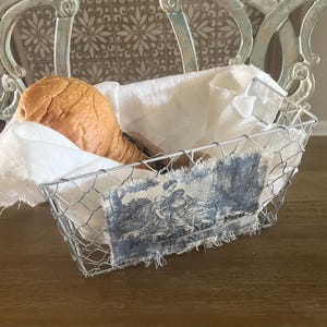 May include: A wire basket with a white cloth lining, holding a loaf of bread. The basket has a decorative blue and white fabric patch with a French scene. The basket is on a wooden surface.