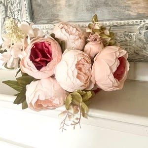 May include: A bouquet of pink and blush peonies with green leaves. The peonies are arranged in a cluster and have a soft, romantic look.