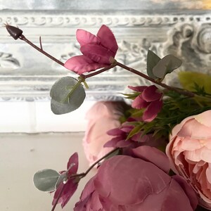 May include: Close-up of artificial flowers in shades of pink and green. The arrangement features large, open peonies and smaller, closed buds. The leaves are a muted green, and the stems are brown. The background is a white, ornate frame.