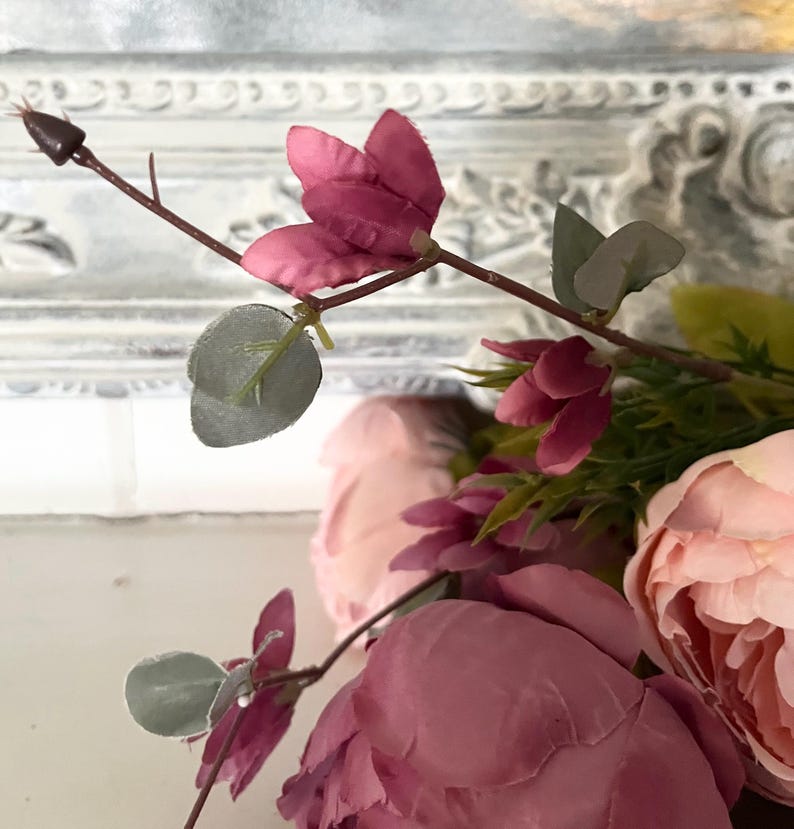 May include: Close-up of artificial flowers in shades of pink and green. The arrangement features large, open peonies and smaller, closed buds. The leaves are a muted green, and the stems are brown. The background is a white, ornate frame.