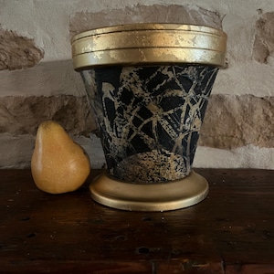 May include: A decorative black and gold vase with a textured pattern and a gold rim and base. A yellow pear sits next to the vase on a dark wooden surface. The vase is a unique home decor item.
