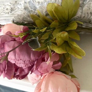 May include: Close-up of a floral arrangement featuring pink and peach-colored peonies and green leaves. The flowers are arranged against a white background with a decorative, carved design.