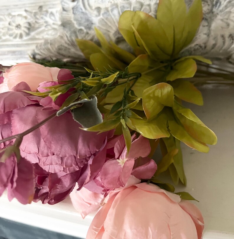 May include: Close-up of a floral arrangement featuring pink and peach-colored peonies and green leaves. The flowers are arranged against a white background with a decorative, carved design.
