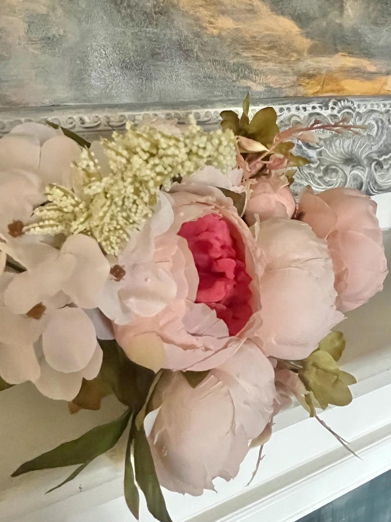 May include: A bouquet of pink and white silk flowers with green leaves. The flowers are arranged in a vase and are sitting on a white surface.