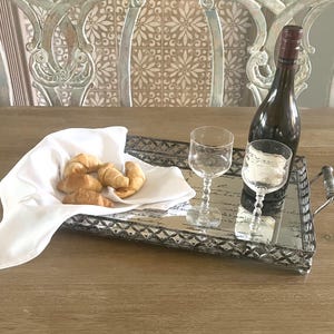 May include: A silver mirrored serving tray with a decorative border and a handwritten inscription. The tray is filled with croissants and two wine glasses. A bottle of wine is on the tray.