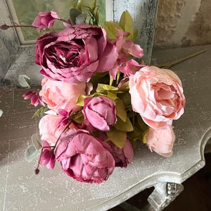 May include: A bouquet of artificial peonies in varying shades of pink and burgundy. The arrangement includes large, fully bloomed flowers and smaller buds, with green leaves and stems. The flowers are arranged on a decorative, light-colored surface.