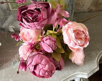 May include: A bouquet of artificial peonies in varying shades of pink and burgundy. The arrangement includes large, fully bloomed flowers and smaller buds, with green leaves and stems. The flowers are arranged on a decorative, light-colored surface.