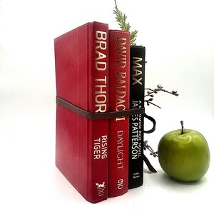May include: A decorative book stack held together with a brown leather strap. The stack includes three books with gold lettering and a sprig of greenery. A green apple sits to the right of the books.