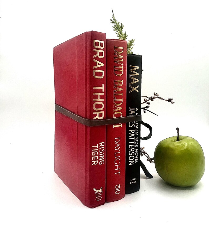 May include: A decorative book stack held together with a brown leather strap. The stack includes three books with gold lettering and a sprig of greenery. A green apple sits to the right of the books.