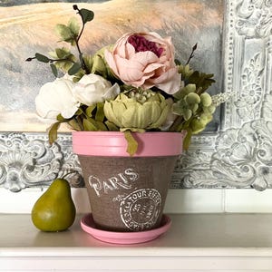 May include: A decorative flower pot with a pink rim and saucer, filled with faux flowers in shades of pink, white, and green. The pot is brown with the word "Paris" and a stamp design. A green pear sits to the left of the pot.
