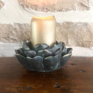 May include: A gold-colored pillar candle sits in a dark teal, flower-shaped candle holder. The candle holder has a metallic finish and is placed on a wooden surface. The background features a textured stone wall.