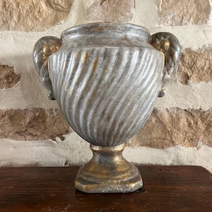 May include: A silver and gold-toned decorative urn with a pedestal base and two side handles. The urn has a textured, swirling pattern and is displayed on a dark wooden surface. The background is a stone wall.