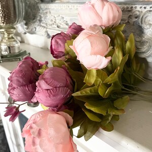 May include: A bouquet of artificial peonies in shades of pink and purple, with green leaves. The flowers are arranged on a white surface, with a decorative mirror in the background. The peonies are in full bloom, showcasing their delicate petals.
