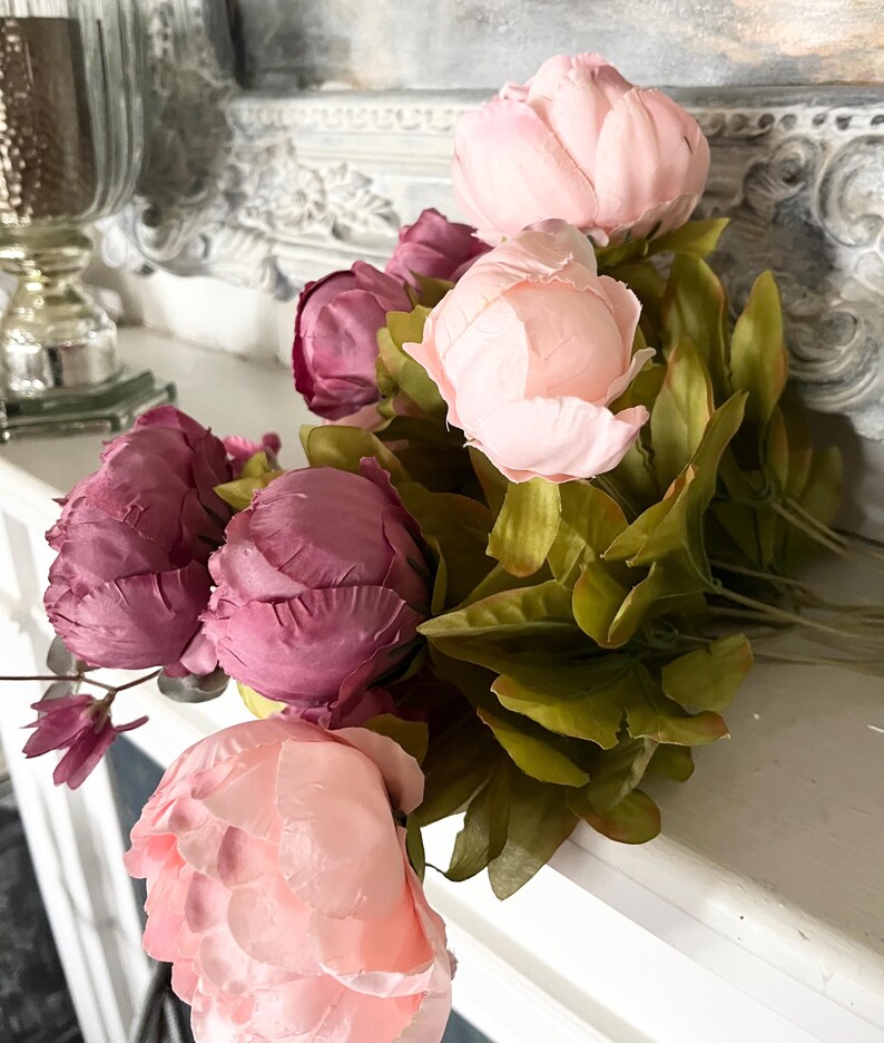 May include: A bouquet of artificial peonies in shades of pink and purple, with green leaves. The flowers are arranged on a white surface, with a decorative mirror in the background. The peonies are in full bloom, showcasing their delicate petals.