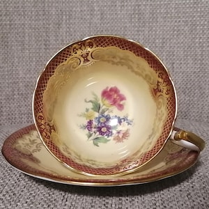 Puede incluir: Un juego de taza y platillo de té vintage. La taza y el platillo son de color crema con un borde granate y dorado. La taza presenta un diseño floral con flores rosas, azules y moradas. El asa es dorada.