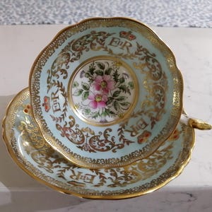 May include: A vintage teacup and saucer set with a light blue background and gold trim. The set features a floral design and the initials "E.R." with a crown.