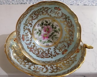 Könnte beinhalten: Ein Vintage-Teetassen- und Untertassen-Set mit hellblauem Hintergrund und goldener Verzierung. Das Set zeigt ein Blumenmuster und die Initialen "E.R." mit einer Krone.