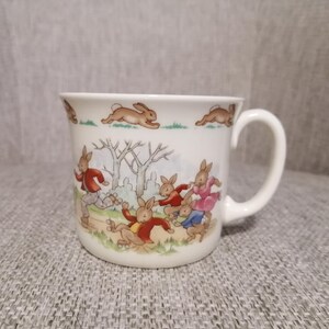 May include: A white ceramic mug with a handle features a colorful illustration of bunnies playing in a forest. The bunnies are wearing red, blue, and pink clothing.
