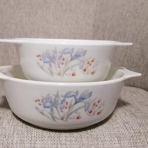 May include: Two white ceramic bowls with a floral design. The bowls are nested together and feature a delicate pattern of blue and purple flowers with green leaves.