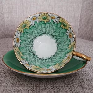 May include: A vintage teacup and saucer set with a green and white floral design. The cup features a white centre with a green and white floral border. The saucer is a solid green colour.