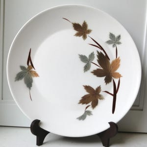 May include: White ceramic plate with a brown and tan leaf design. The leaves are arranged on brown stems. The plate is displayed on a dark brown wooden stand. A decorative plate for serving.