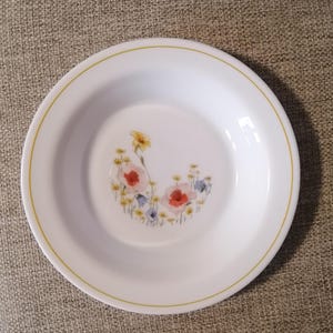 May include: A white ceramic bowl with a yellow rim and a floral design featuring pink, red, and blue flowers. The bowl is perfect for serving soup, cereal, or snacks.