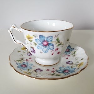May include: A white teacup and saucer set with a floral pattern. The cup and saucer have a gold trim and feature blue, yellow, pink, and green flowers.