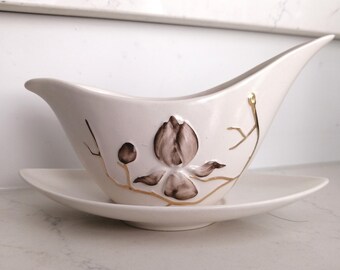 May include: Cream-colored ceramic gravy boat with a matching saucer. The boat features a floral design in brown and gold. The gravy boat has a unique, elongated shape with a pointed spout.
