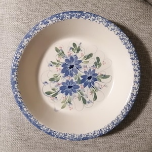 Könnte beinhalten: Eine weiße Keramik-Tortenplatte mit einem blau-weiß gesprenkelten Rand. Die Mitte der Platte zeigt ein handgemaltes Design aus blauen Blumen mit grünen Blättern und rosa Akzenten.