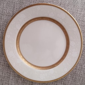 Puede incluir: Un plato de cerámica blanco con un borde dorado y un delicado patrón floral. El plato tiene un fondo blanco con un borde dorado y un delicado patrón floral.