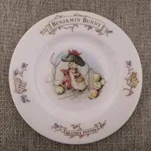 May include: A white ceramic plate featuring a colourful illustration of Benjamin Bunny from Beatrix Potter's children's book. The plate has a blue and white ribbon with the text "Benjamin Bunny" and a green ribbon with the text "Beatrix Potter".