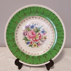 May include: A white ceramic dinner plate with a green and gold border. The center of the plate features a floral design with pink, yellow, and blue flowers.