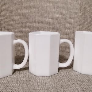 May include: Three white ceramic mugs with octagonal sides and handles. The mugs are arranged in a row on a textured surface.