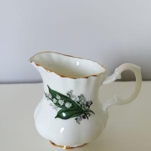 Op de afbeelding: Een witte porseleinen roomkan met een gouden rand en een bloemenontwerp met lelietjes-van-dalen.