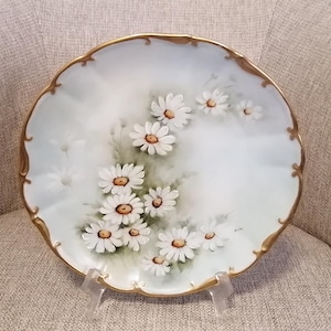 Könnte beinhalten: Ein weißer Porzellanteller mit goldfarbenem Rand und einem handgemalten Design aus weißen Gänseblümchen auf grünem Hintergrund.