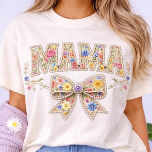 Könnte beinhalten: Cremefarbenes T-Shirt mit dem Wort "MAMA" in floralem Design. Darunter eine florale Schleife mit Perlenakzenten. Das Shirt hat einen Rundhalsausschnitt und kurze Ärmel.