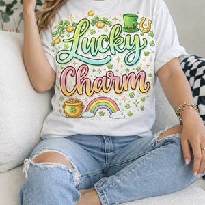 May include: White t-shirt with a colorful "Lucky Charm" graphic. The design includes a rainbow, pot of gold, shamrocks, and a green hat. The shirt is paired with light blue jeans.