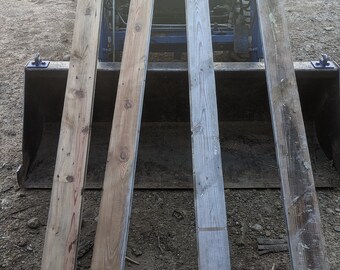 May include: Four weathered wooden planks, likely reclaimed lumber, are stacked on top of a black metal tractor bucket. The planks are various shades of brown and gray, showing signs of age and use.