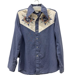 May include: A long-sleeved blue button-down shirt featuring a cream-colored quilted yoke across the chest and shoulders. The yoke is decorated with two star-shaped appliqués in shades of blue, green, and floral patterns with a central dark circle.