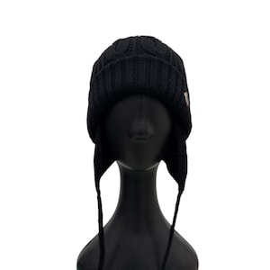 May include: A black knit beanie featuring a cable knit pattern on the crown, a ribbed cuff, and two side ear flaps with long, thin braided ties hanging down. The hat is displayed on a black mannequin head, showcasing its textured construction.