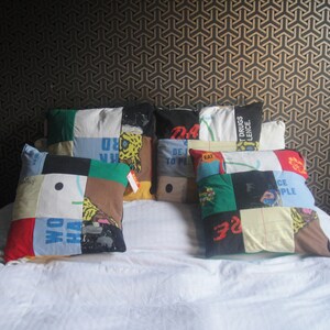 May include: A white bed with six colorful patchwork pillows. The pillows are made from different fabrics and have various text and designs. The pillows are arranged in a row on the bed.