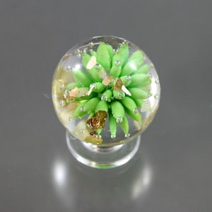 May include: A clear glass sphere with a green flower-like design inside. The sphere is filled with small air bubbles and gold flecks. The sphere is mounted on a clear glass base.